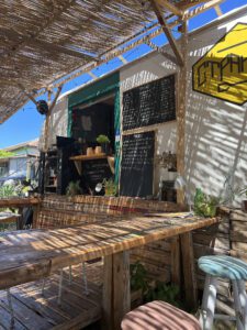 Strandja Food Truck-tilausluukku, ruokalistat kirjoitettuna käsin liitutaululle, keltainen logo vaunun kyljessä ja puiset tuolit. Valo siivilöityy ruokokaton lävitse.
